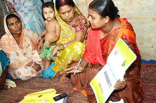 29244770927_6b3aa1cc7f_z A woman wears a bright orange saree and holds up a poster with the JEEViKA number on it. She has a mobile phone placed near her. A group of women, one with a child, are listening to her as she explains how to use JEEViKA Vaani